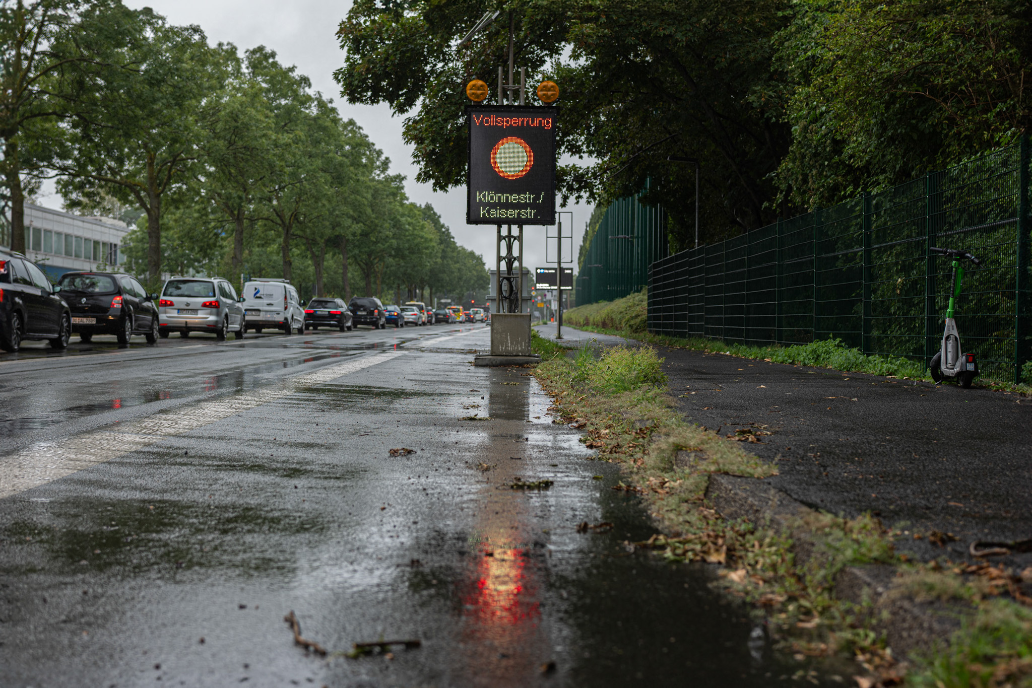 Verkehrszeichen mit der Aufschrift 'Vollsperrung' an einer nassen Straße, umgeben von im Stau stehenden Autos und einem E-Scooter.