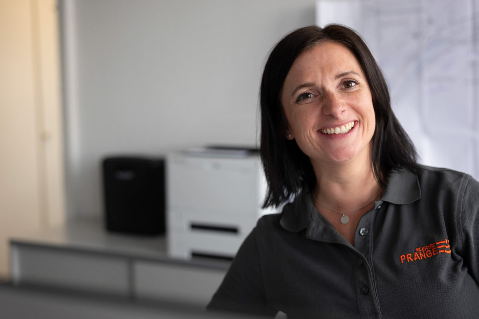 Frau mit schwarzem Haar und grauem Poloshirt, die lächelnd am Schreibtisch sitzt. Im Hintergrund sind Bürogeräte sichtbar.