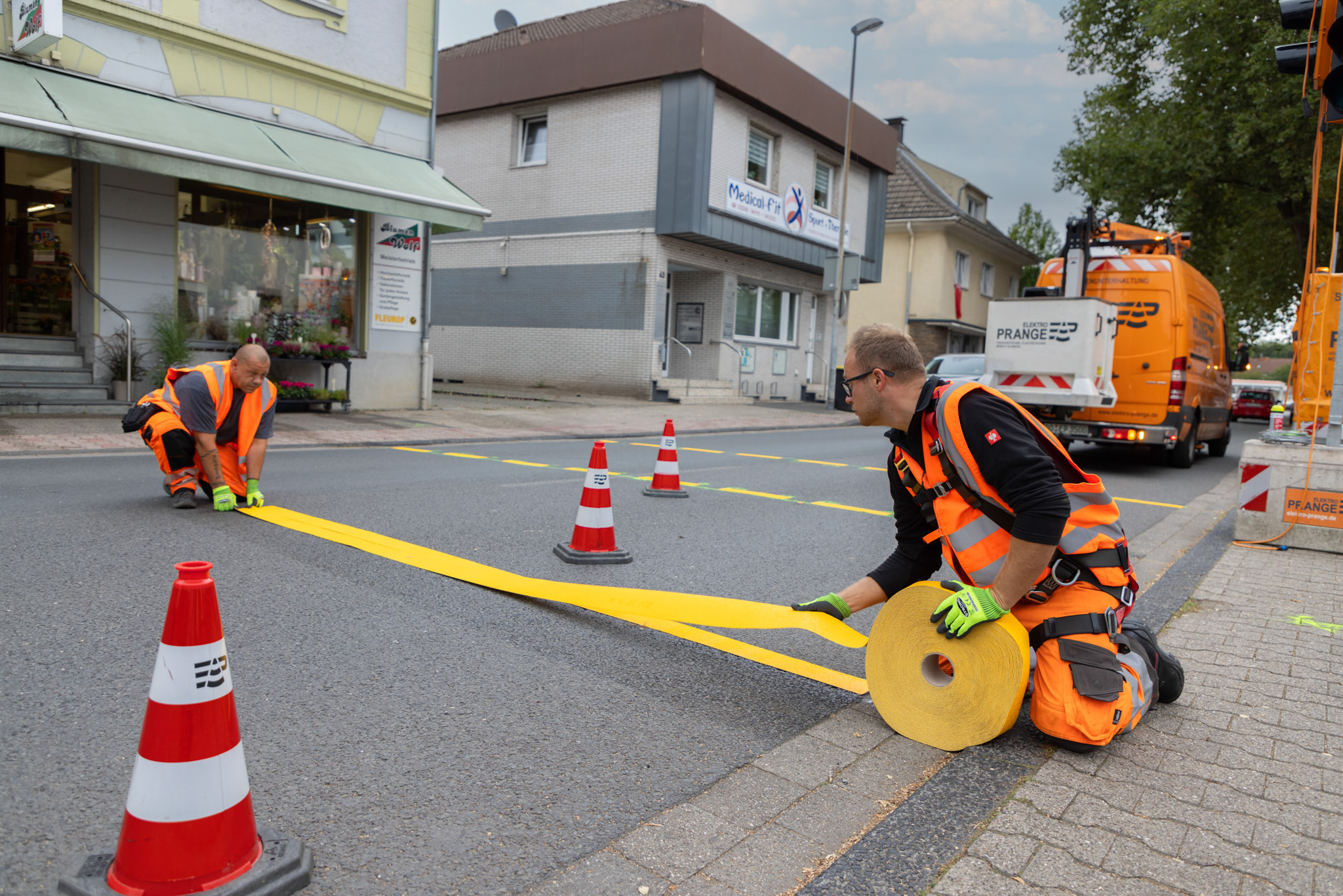 Zwei Arbeiter in orangefarbener Sicherheitskleidung markieren eine Straße mit gelbem Farbband und roten Verkehrshütchen.