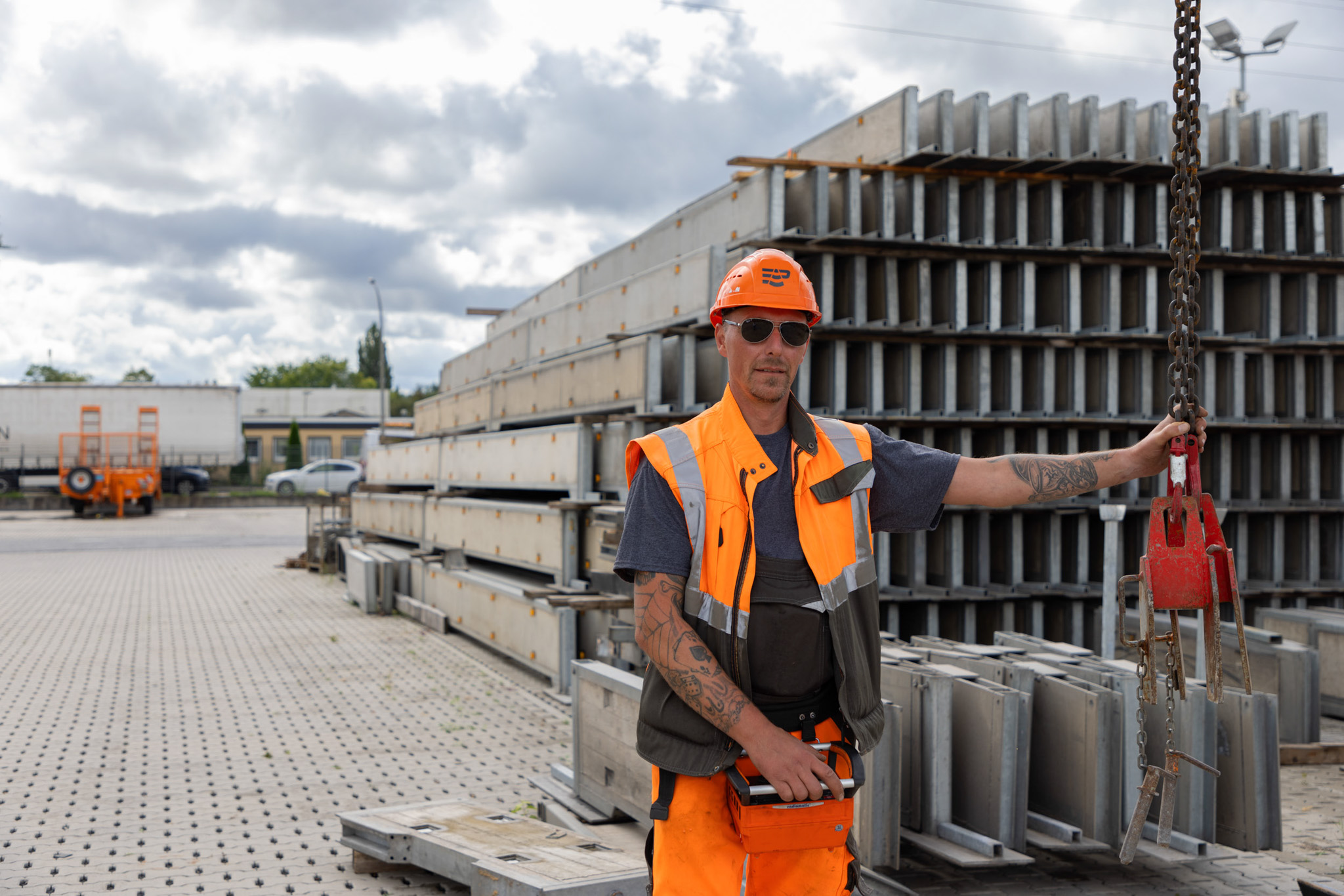 Arbeiter in orangefarbener Sicherheitskleidung mit Helm, der einen Kranhaken in einer Baustellenumgebung hält.