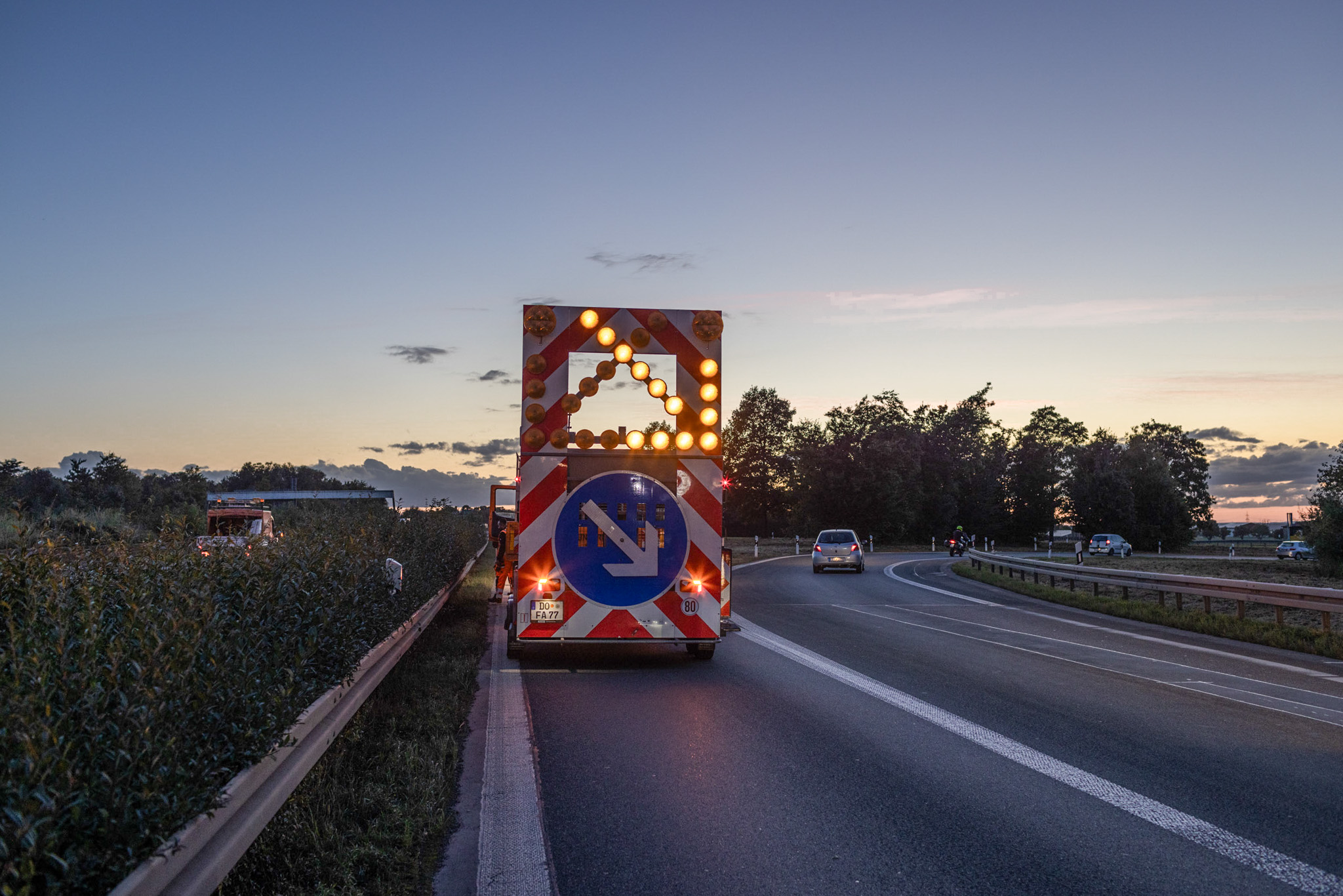 Rückansicht eines Fahrzeugs mit Warnlichtern und Verkehrssymbol auf einer Straße bei Sonnenuntergang.