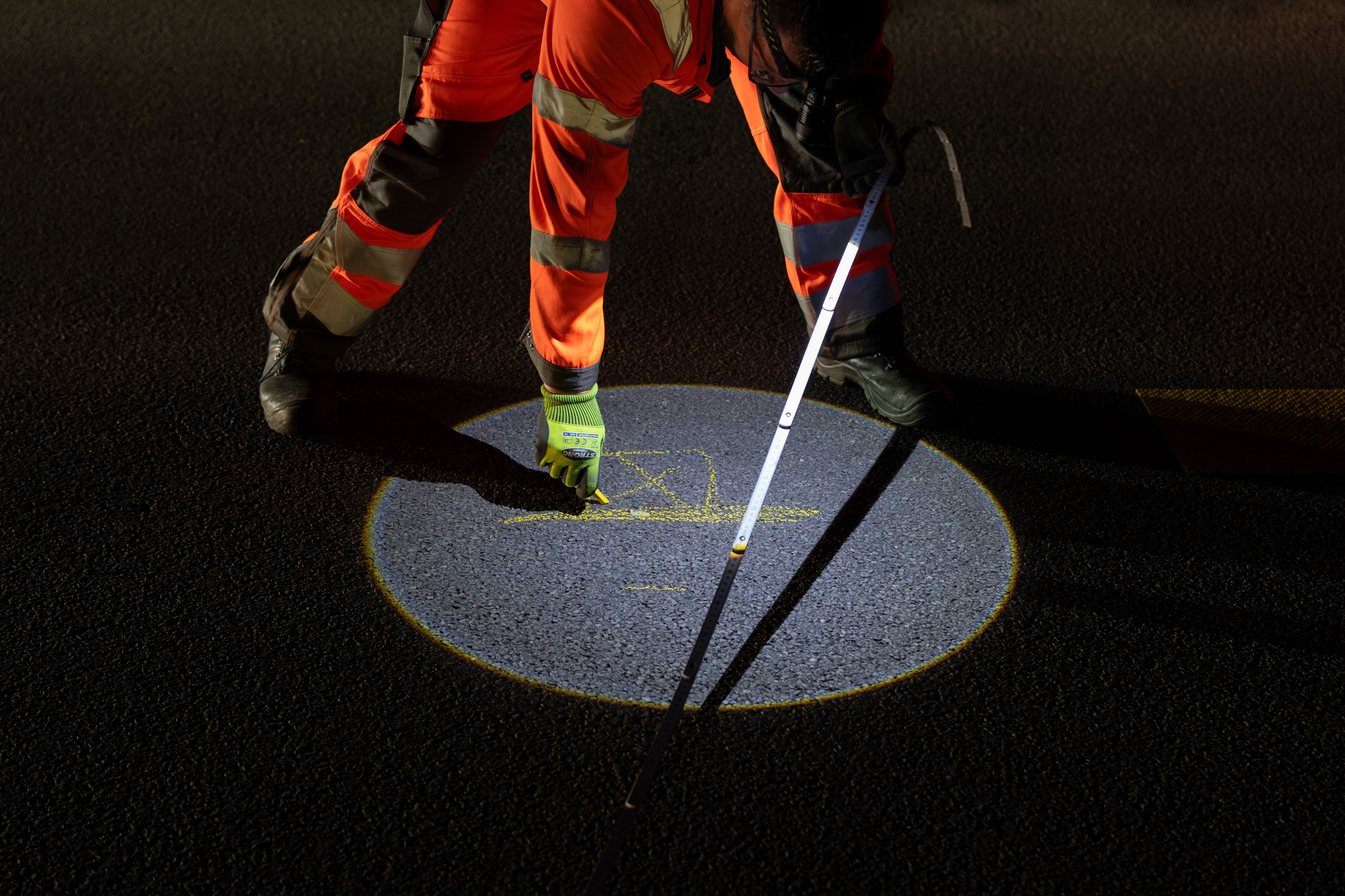 Eine Person markiert mit gelber Kreide einen Punkt auf einer Straße.