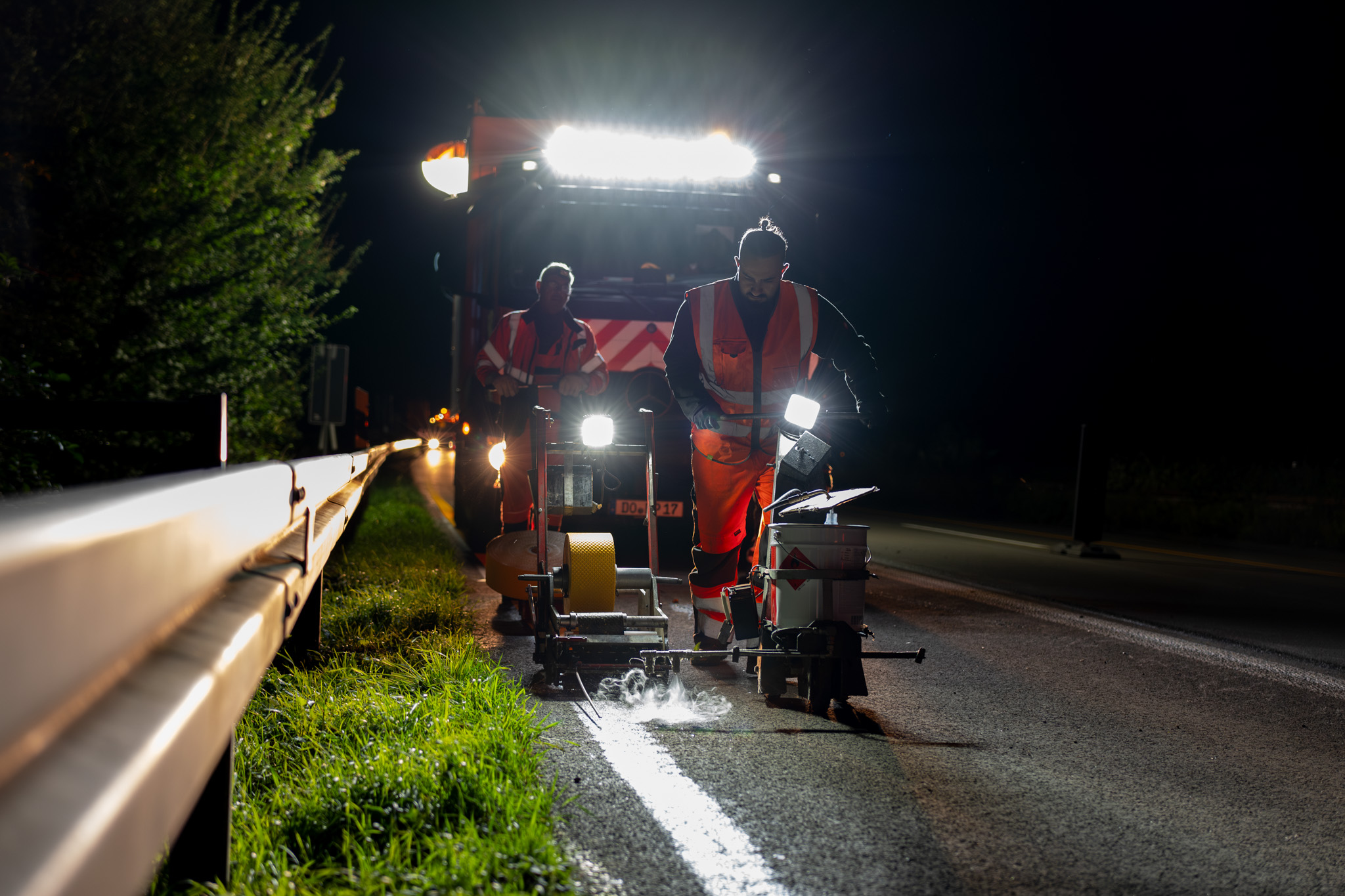 Zwei Menschen bringen nachts auf der Autobahn temporäre Fahrbahnmarkierungen an. Hinter ihnen ein LKW, der alles ausleuchtet.