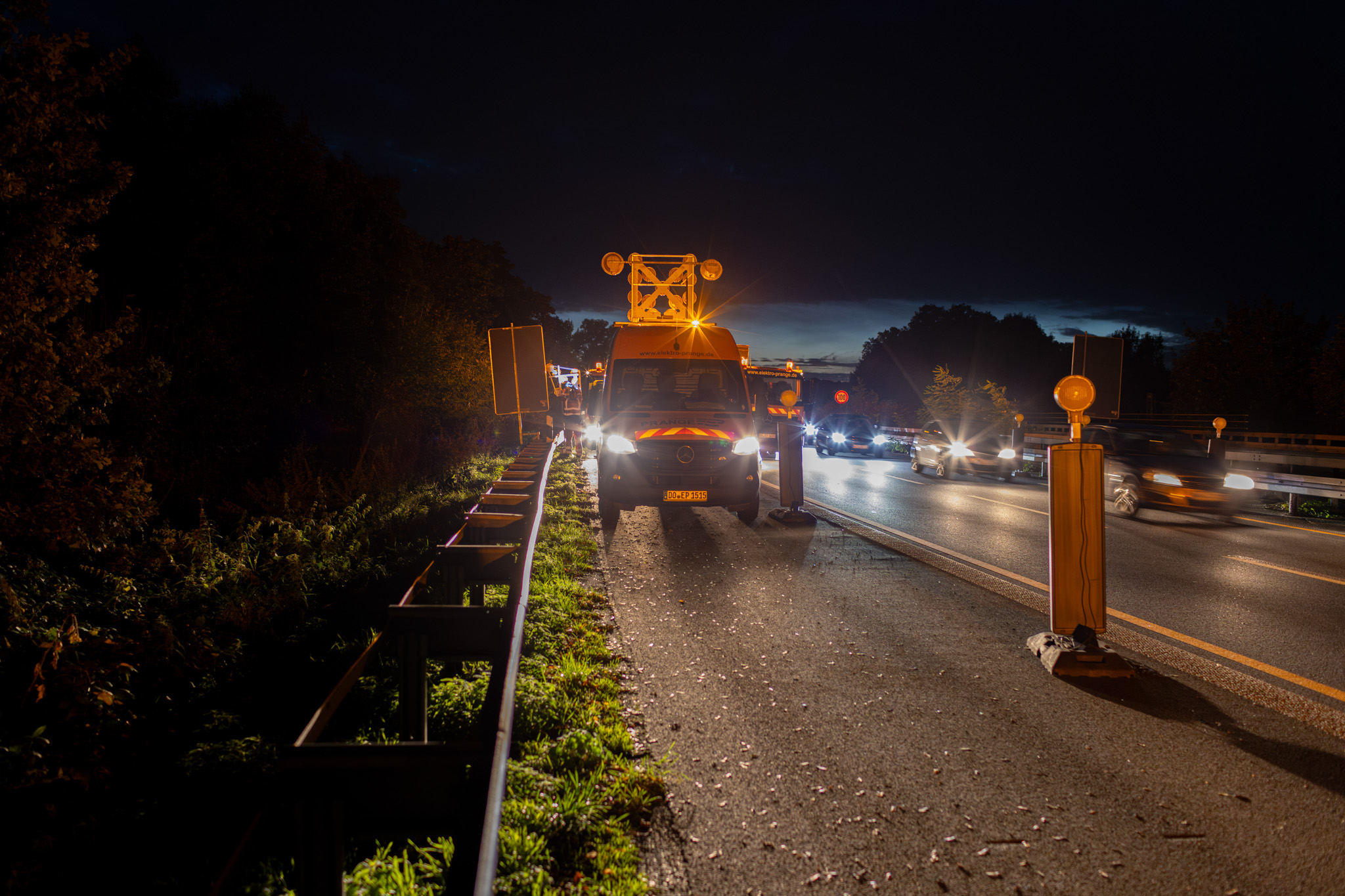 Baufahrzeug mit Warnlichtern auf einer Autobahn bei Nacht, umgeben von Verkehr und Baustellenabsperrungen.