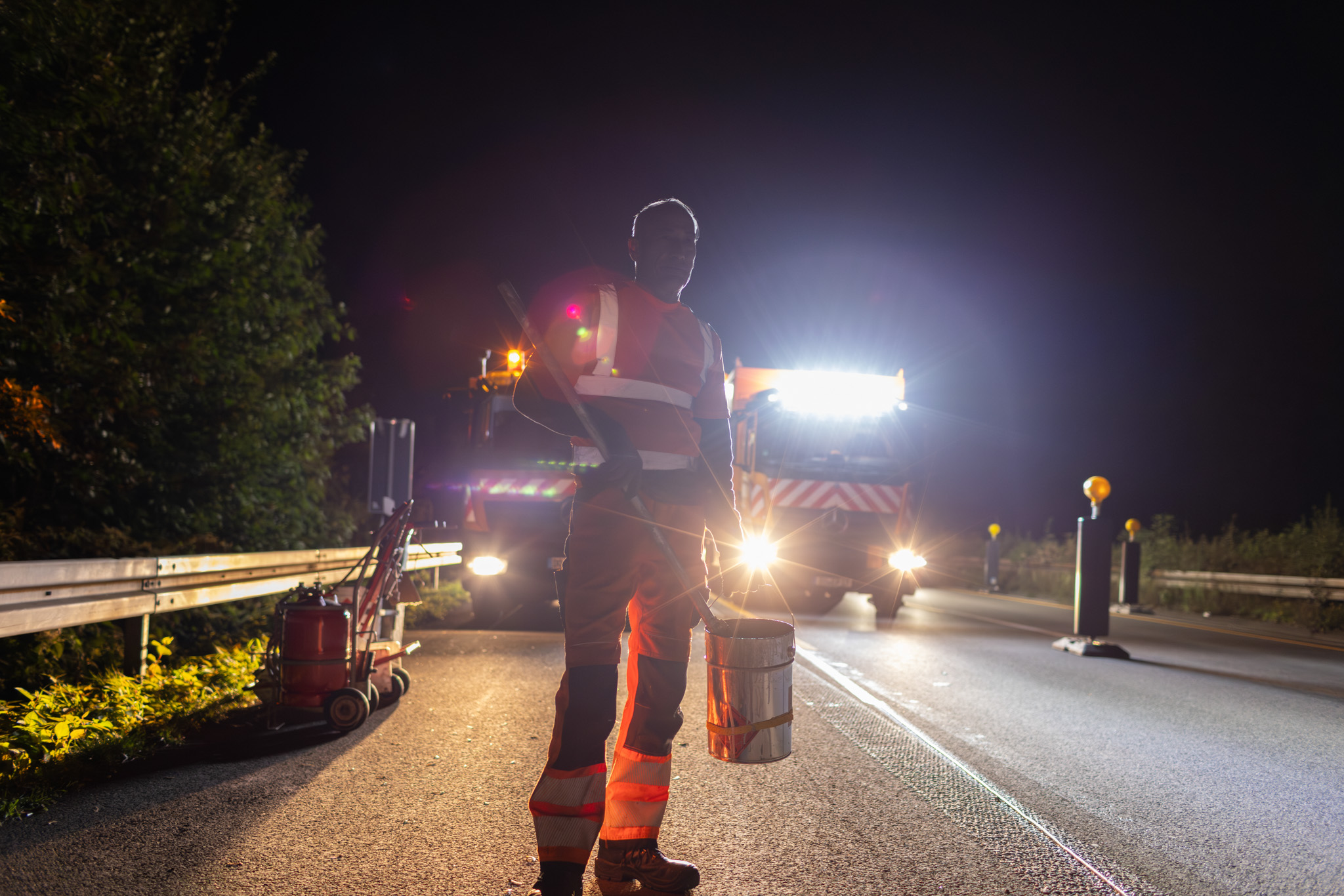 Ein Monteur für Verkehrssicherung steht nachts auf der Autobahn mit einem Eimer in der Hand. Hinter ihm stehen zwei LKWs mit orangen Warnleuchten.