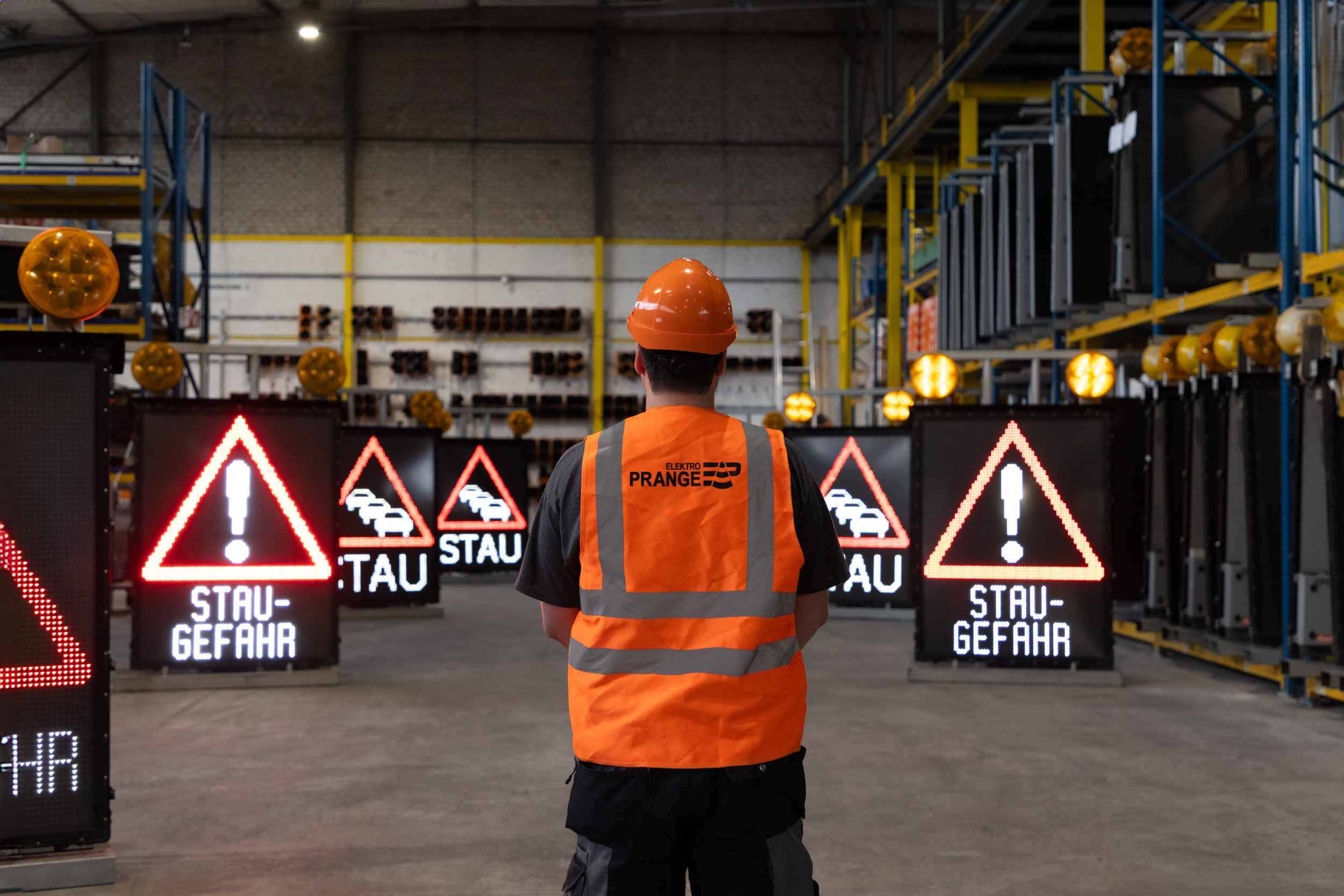 Mitarbeiter in orangefarbener Warnweste mit Helm steht vor digitalen Verkehrshinweis-Displays in einem Lager.